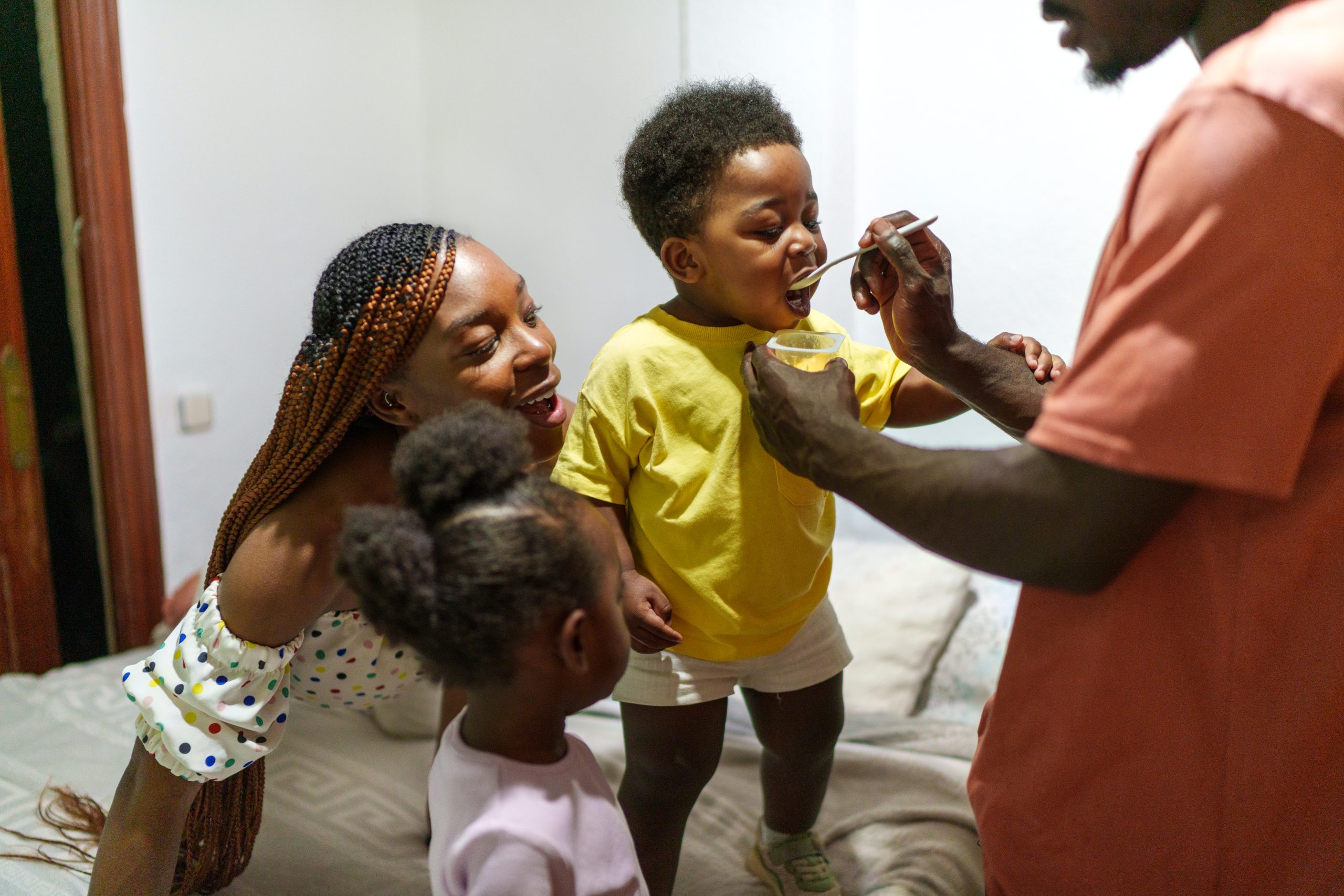 Father Shares Yogurt with Smiling Children in a Cozy Bedroom Setting