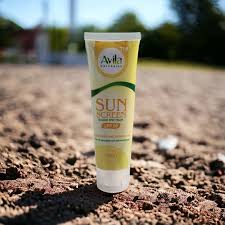 A tube of avila sunscreen stands on a rough, dry ground with greenery visible in the blurred background.