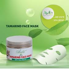 A jar of avila tamarind face mask next to a facial mask sheet, displayed against a green background with water droplets and text promoting the product.