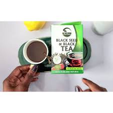 Hands holding a cup of tea next to a box of black seed tea on a table with a lemon slice and a spoon.