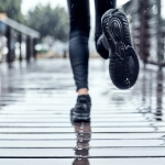 Close-up of a person running on a wet wooden bridge. The ground is reflective from the rain, and the runner's black running shoes and legs are partially visible.