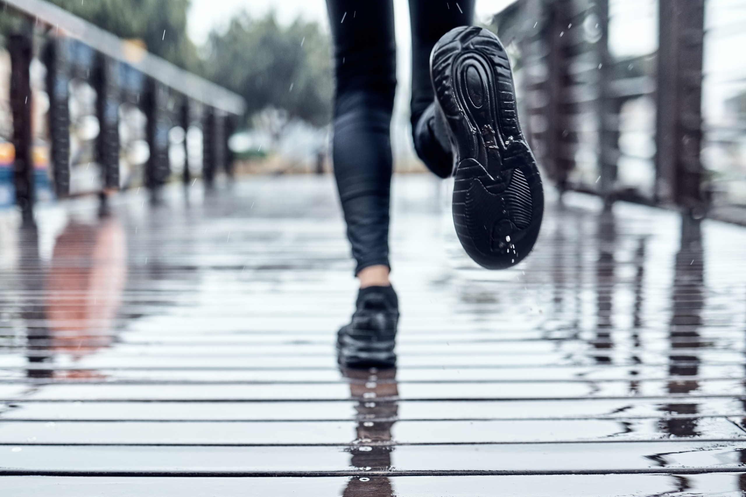 Close-up of a person running on a wet wooden bridge. The ground is reflective from the rain, and the runner's black running shoes and legs are partially visible.